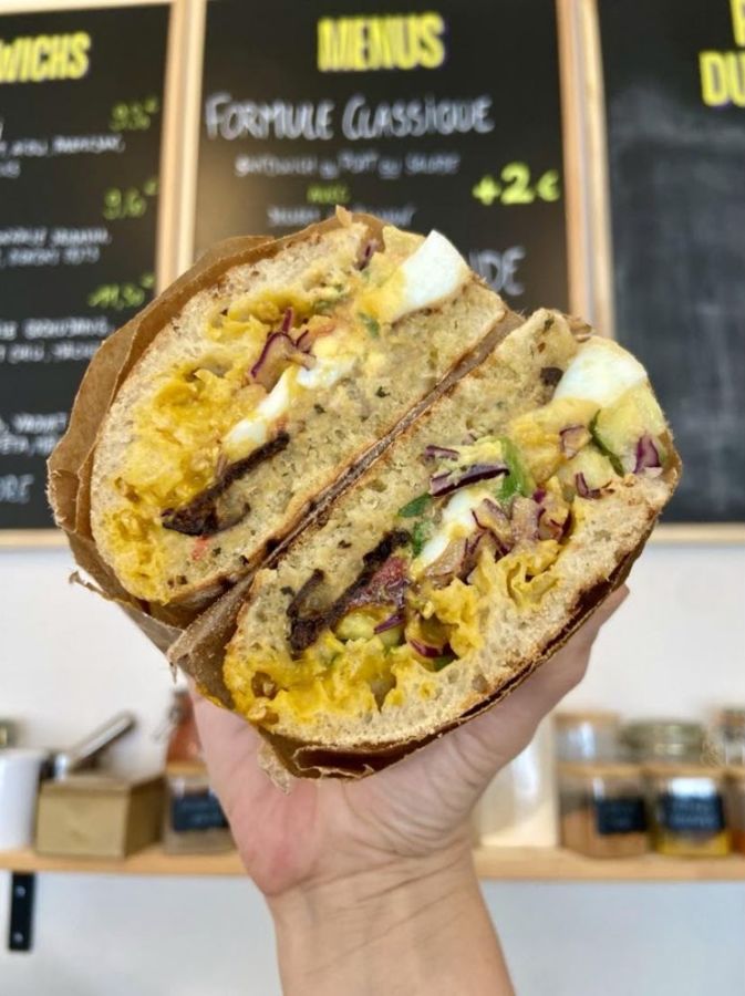 Sandwich ciabatta coupé en deux, tenu à la main devant les ardoises du menu